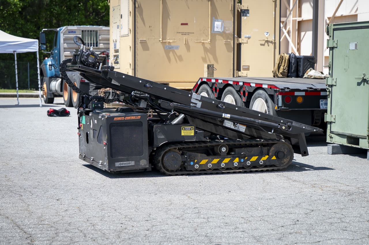 MPEP with its deck implement lowered beside an Air Force flatbed trailer, hazard chevrons visible on the track guard.